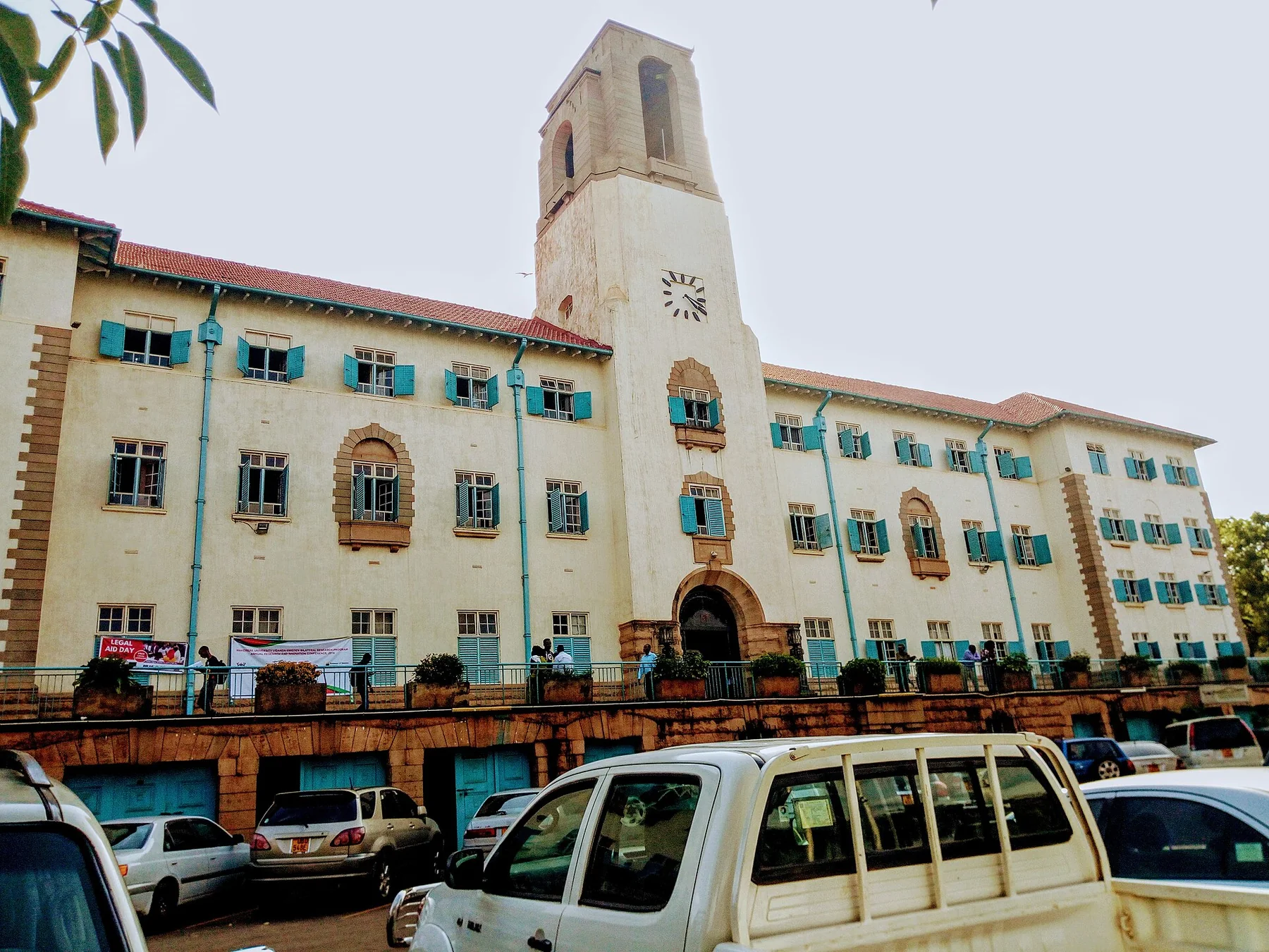 Makerere University'nin uzun krem rengi ana binası park etmiş araçların ardında yükseliyor; merkezde saat kulesi, mavi-yeşil panjurlar, kemerler ve kırmızı kiremitli çatı var.