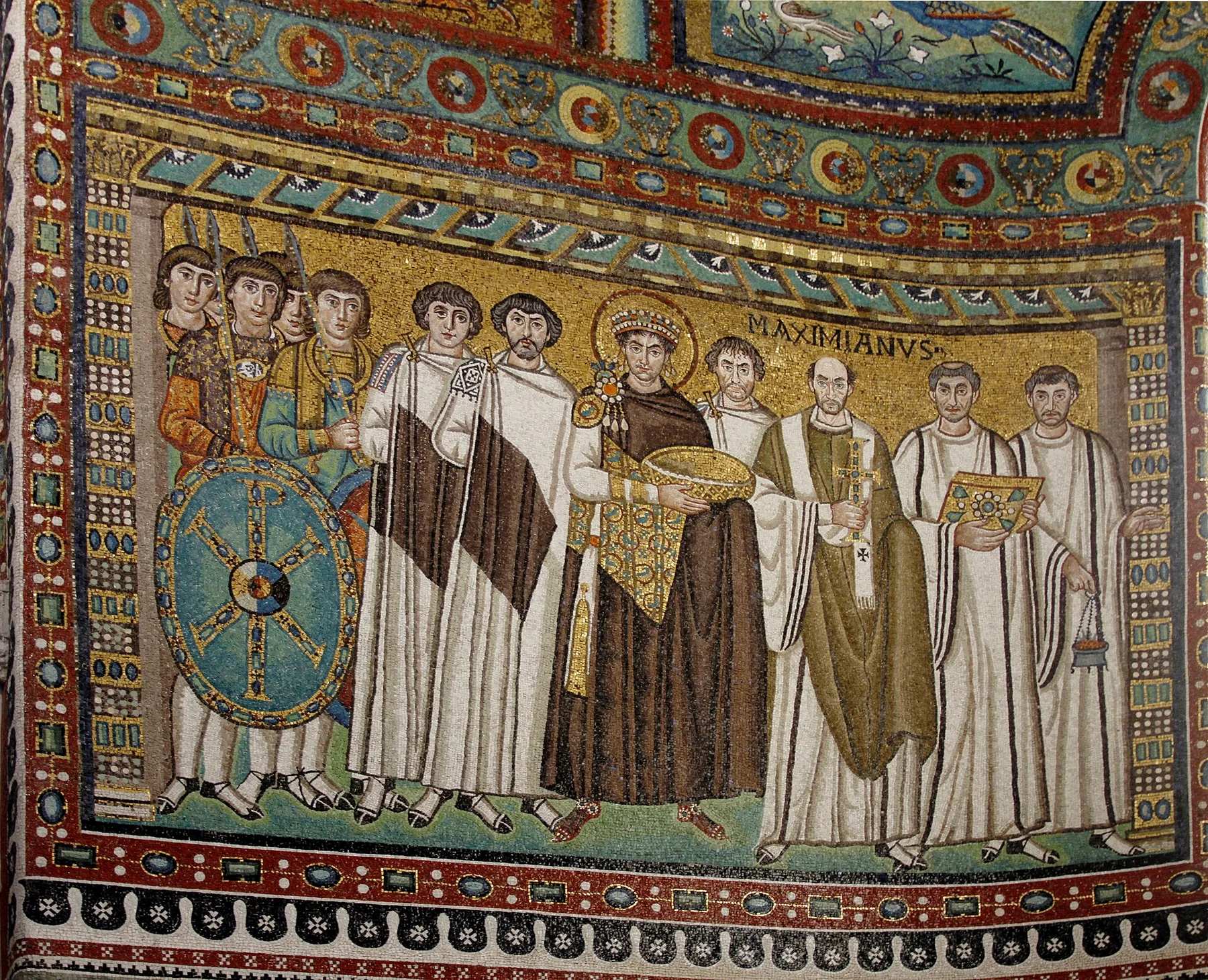 İmparator Iustinianus altın zeminli mozaiğin merkezinde büyük sunu kabı tutuyor; asker, saray görevlisi, Piskopos Maximianus, din görevlisi, haç, İncil kitabı ve buhurdanla çevrili.