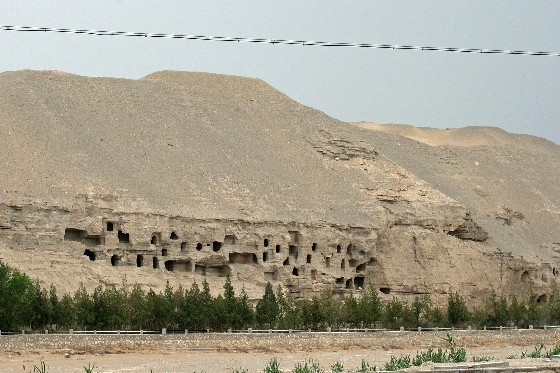 Soluk renkli bir uçuruma oyulmuş koyu mağara açıklıkları ve küçük oda sıraları, Dunhuang yakınındaki çöl peyzajında ağaçların üzerinde yükselir.