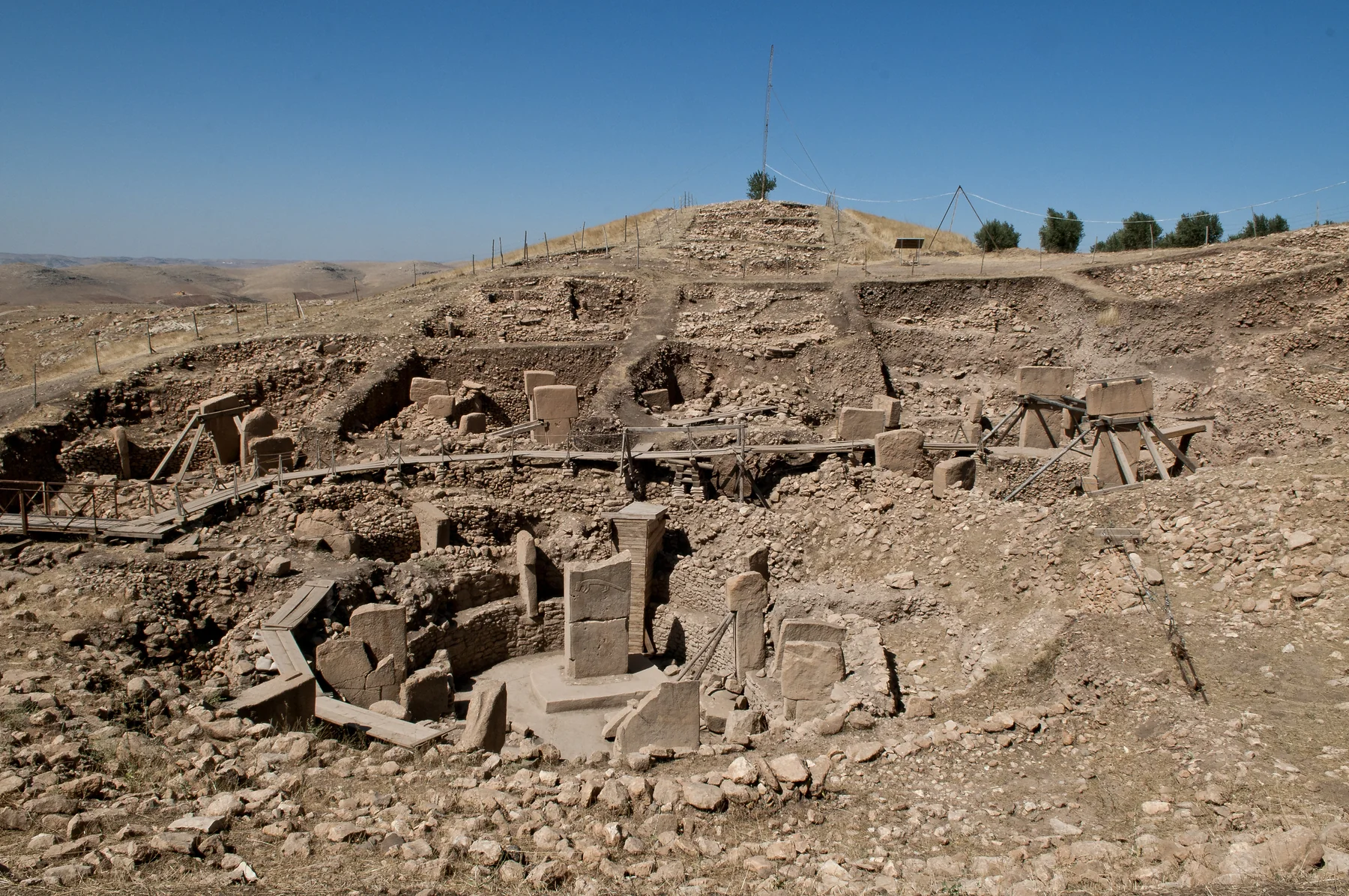 Dik T biçimli kireçtaşı sütunların bulunduğu dairesel taş yapıların kazıldığı tepe; yürüyüş yolları ve modern destekler de görünüyor.