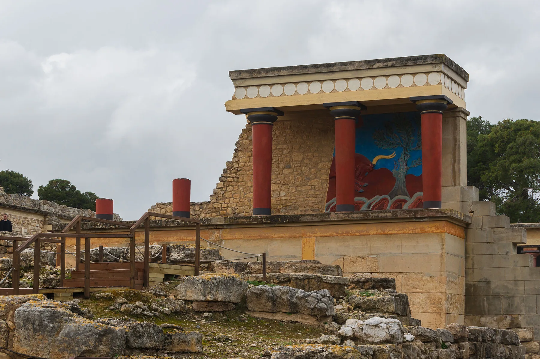 Kırmızı, aşağı doğru incelen sütunlar ve mavi zemin üzerindeki boğa resmiyle yeniden kurulmuş Knossos cephesinin yanında antik taş kalıntıları.