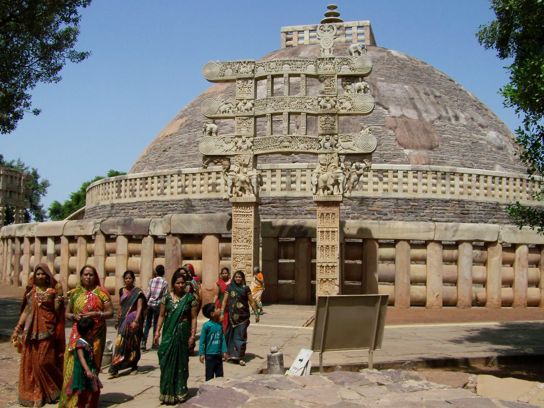 Ziyaretçiler mavi gökyüzü altında Sanchi 1 numaralı Stupa'nın oyma doğu kapısı ve büyük yarım küre kubbesi önünde.