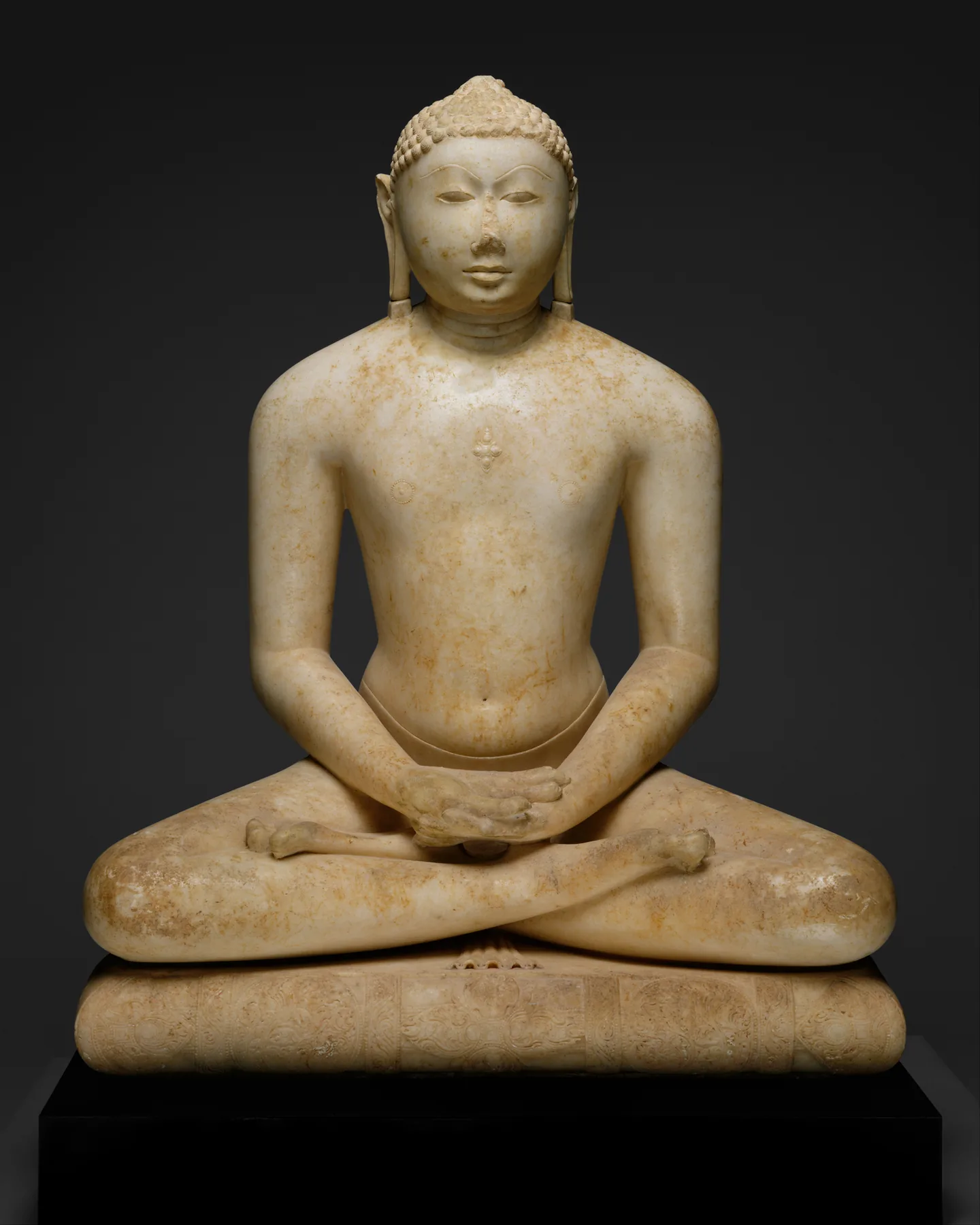 Beyaz mermer Tirthankara bezemeli minder üzerinde dik meditasyonda oturuyor; simetrik bacakları, kucakta dinlenen elleri ve açık, öne bakan gözleri var.