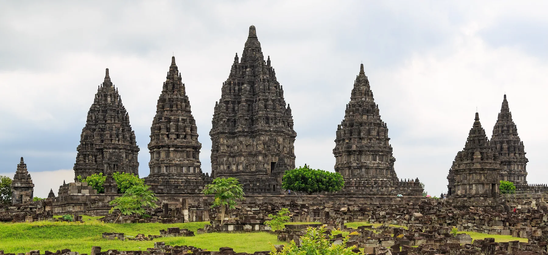 Prambanan tapınak yerleşkesinin yüksek ve kümelenmiş kuleleri yeşil çimenlerin ve dağınık koyu taş kalıntıların üzerinde, bulutlu bir göğe doğru yükselir.