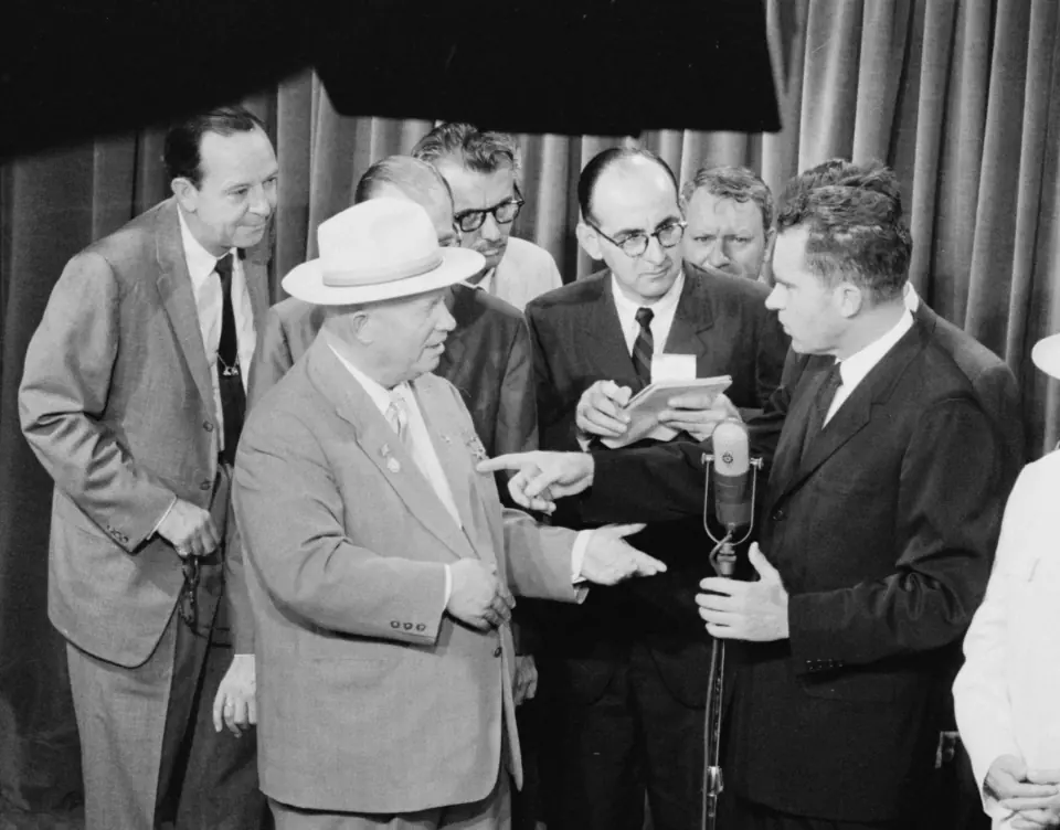 Açık renk takım elbiseli ve şapkalı Nikita Kruşçev, mikrofonun yanındaki Richard Nixon’a doğru el hareketi yaparken çevirmenler, görevliler ve gazeteciler çevrelerini doldurur.