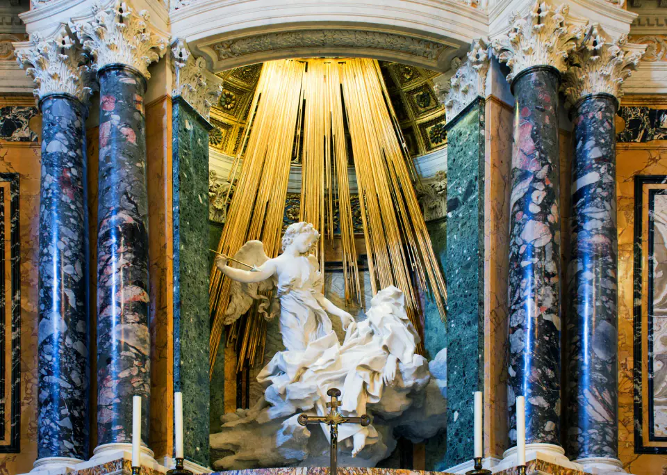 Bernini'nin beyaz mermer Aziz Teresa ve melek figürleri, Cornaro Şapeli'nin renkli mermer sütunları arasında yaldızlı ışınların altında görülüyor.