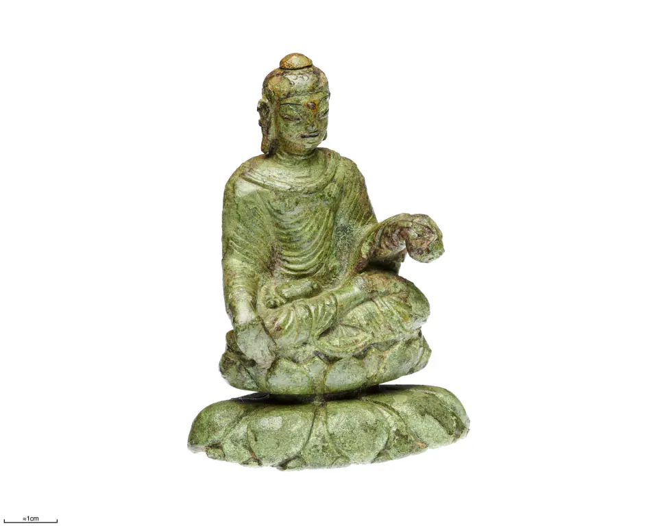 Küçük yeşil bakır alaşımlı Buda beyaz zemin önünde çift lotus tahtında oturuyor; yüzünde altın, gümüş ve pigment izleri var.