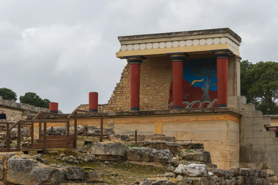 Knossos'ta antik taş kalıntılarının yanında kırmızı sütunlar ve modern bir boğa freski kopyası taşıyan yeniden kurulmuş Kuzey Girişi.