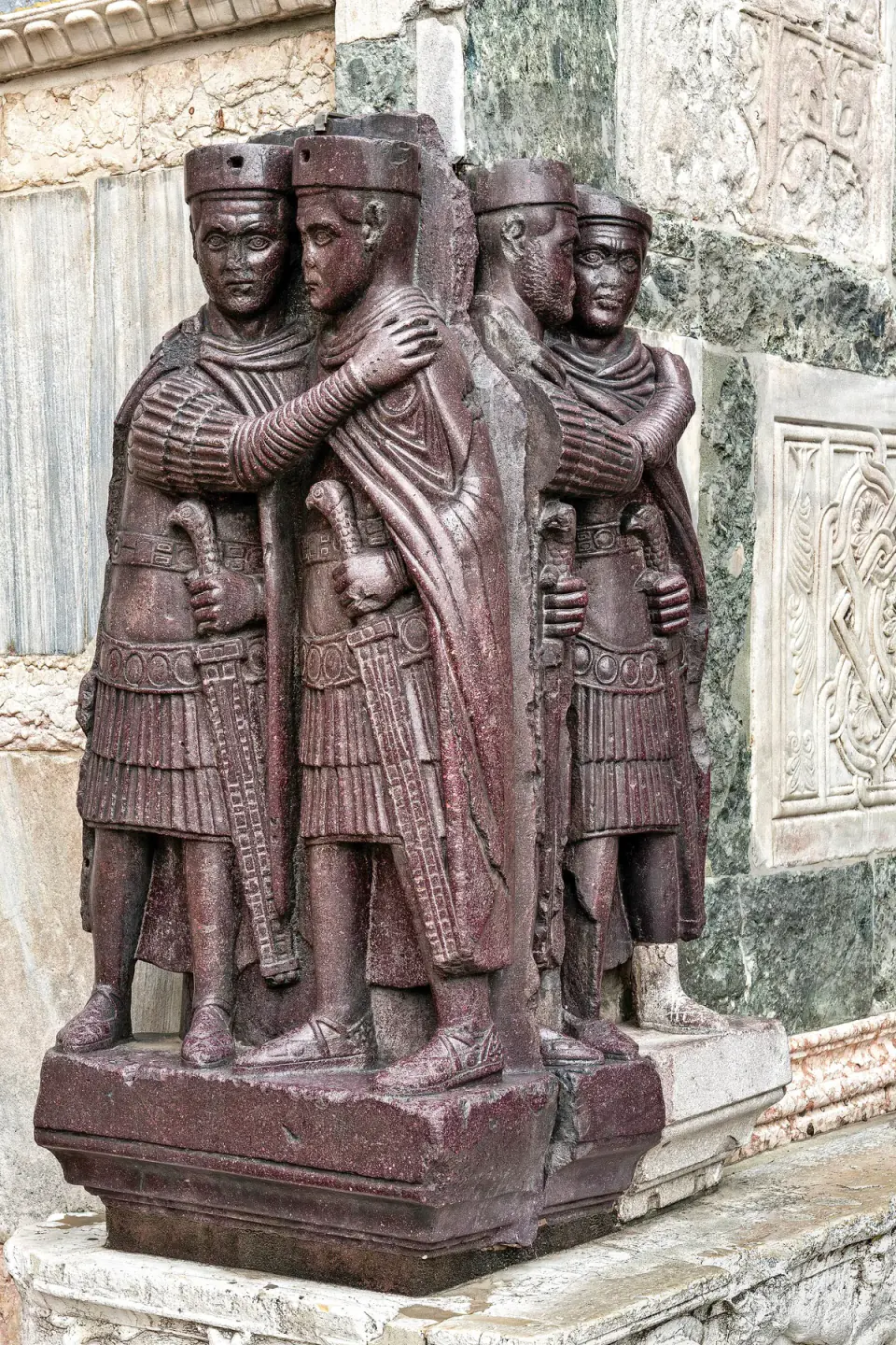 Kırmızı-mor porfirden dört hükümdar, benzer askerî giysilerle ikili gruplar hâlinde birbirine sarılıyor ve San Marco Bazilikası duvarında kılıçlarını tutuyor.