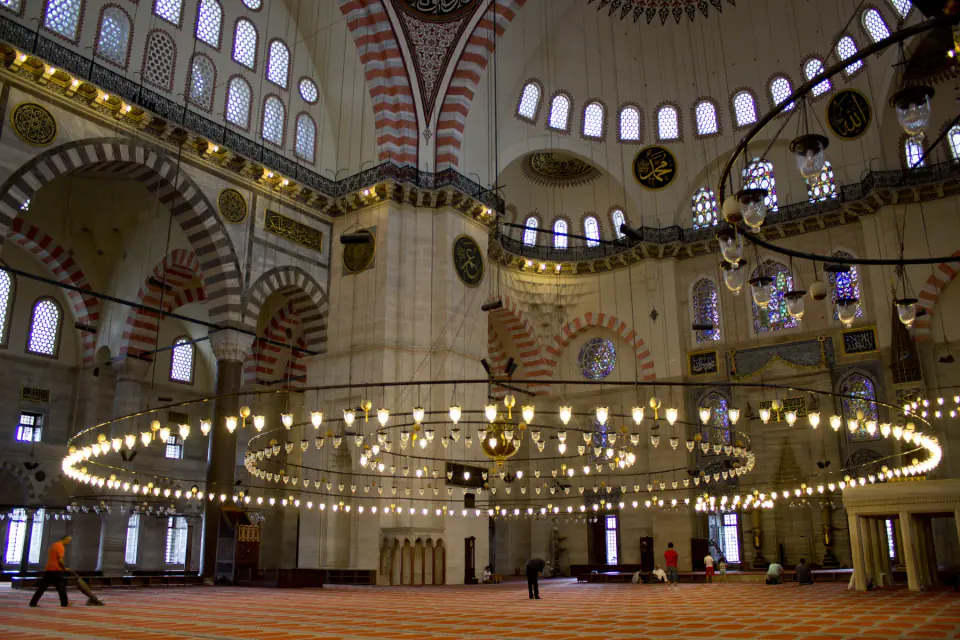 Süleymaniye Camii içindeki geniş görünüşte çizgili kemerler, pencere sıraları, hat levhaları, asılı kandiller ve halılarla kaplı büyük ibadet alanı kubbeli mekânı düzenliyor.