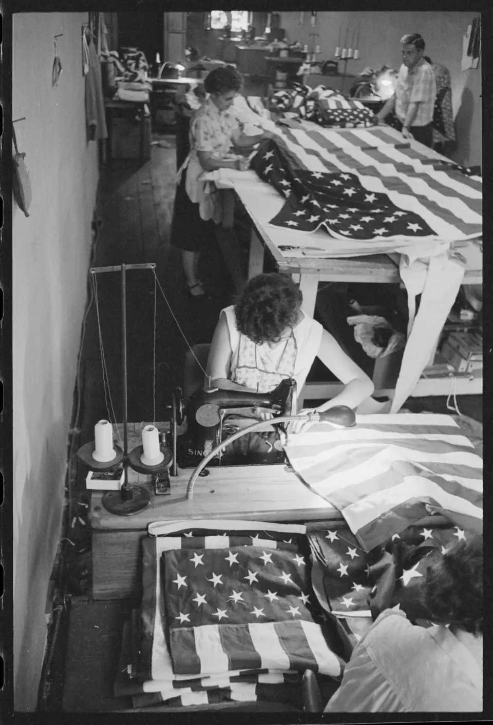Üç kadın farklı çalışma masalarında yinelenen Amerika Birleşik Devletleri bayraklarını dikip düzenlerken bir erkek dar fabrika odasının arkasında durur.