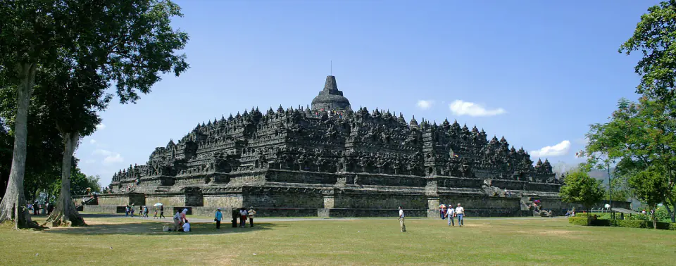 Borobudur’un geniş basamaklı kütlesi yeşil bir düzlükte yükselir; terasları küçük stupalarla doludur ve tepede tek bir merkezî stupa bulunur.